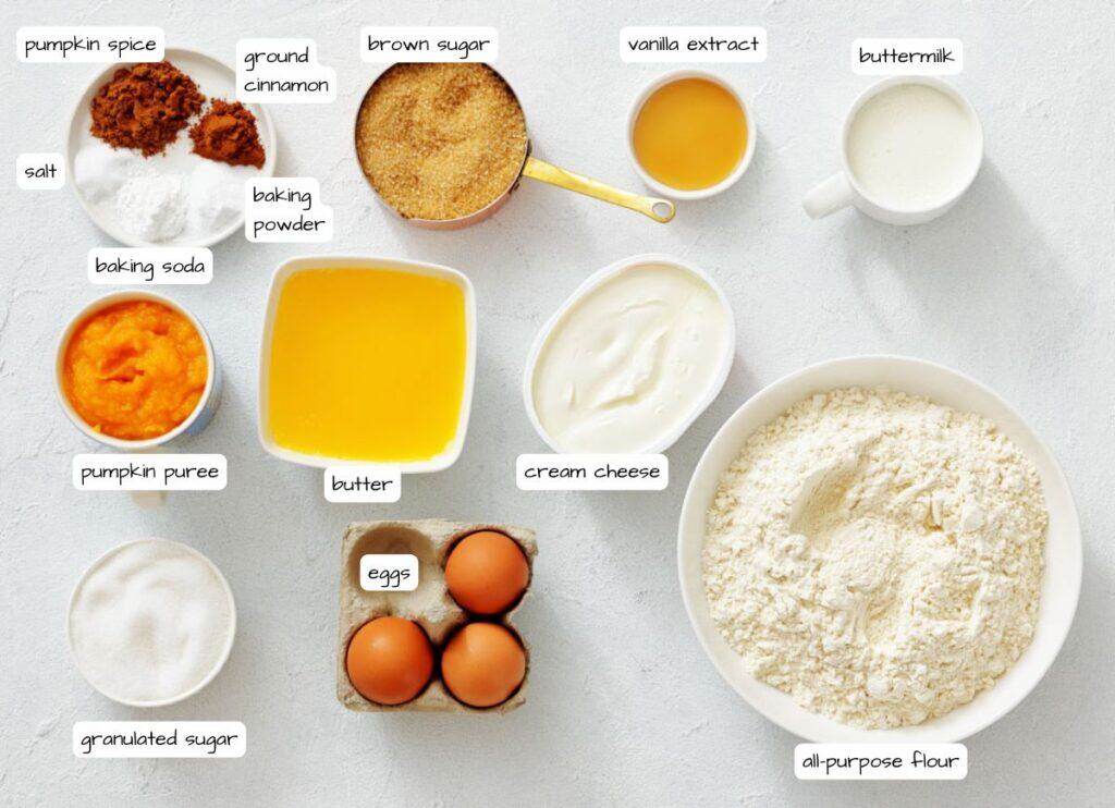 Ingredients for baking are arranged on a white surface, including flour, eggs, butter, pumpkin puree, sugars, spices, cream cheese, baking powder, baking soda, salt, vanilla extract, and buttermilk.