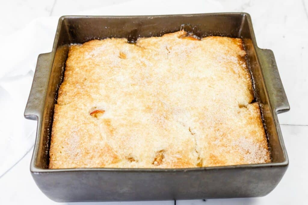 A golden-brown cobbler with a sugar-dusted crust in a square metal baking pan on a white surface.