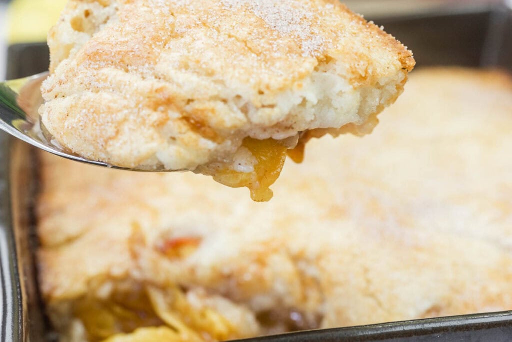 A spoon lifting a serving of peach cobbler with a golden, sugary crust from a baking pan.
