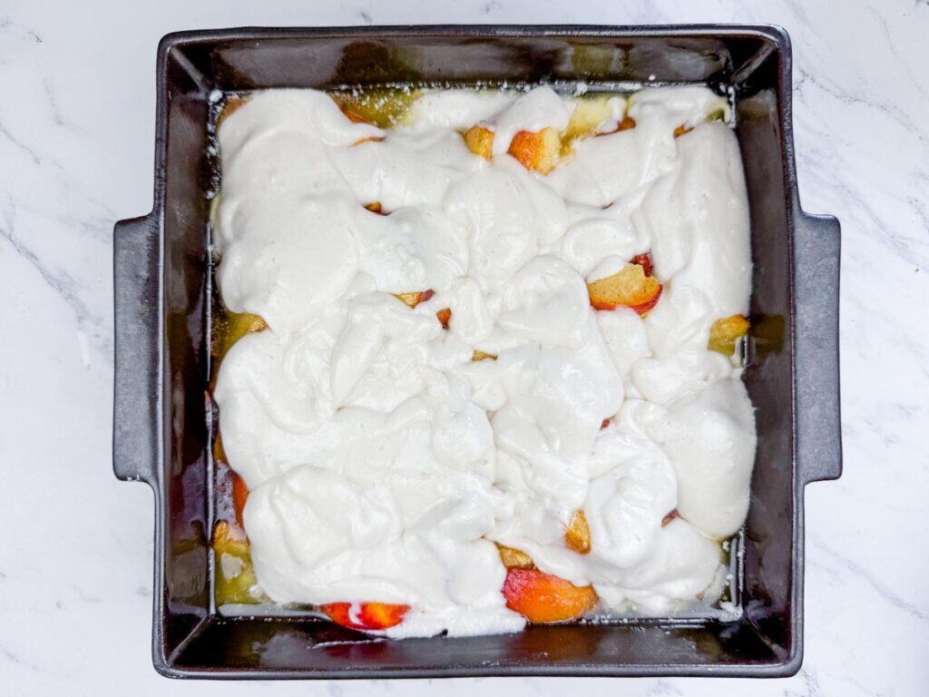 A square baking pan with sliced peaches partially covered by an uncooked white batter, sitting on a white marble surface.