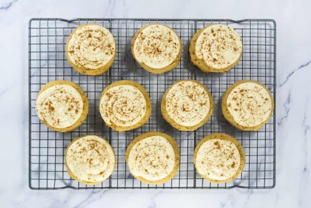 Ten frosted cookies topped with a sprinkle of cinnamon are arranged in rows on a metal cooling rack over a white marble surface.