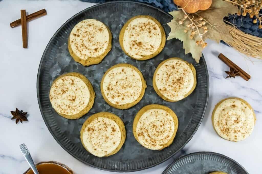 A round metal tray holds eight frosted cookies sprinkled with cinnamon. Nearby are spices, a small bowl of tea, and autumn-themed decorations on a marble surface.