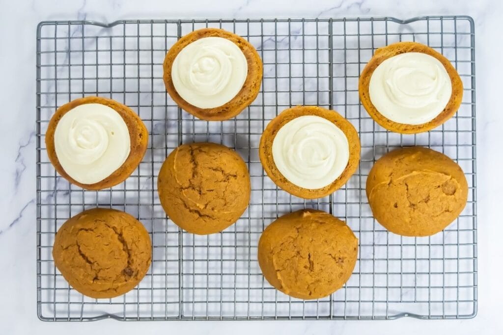 Pumpkin whoopie pie halves on a cooling rack, some topped with cream filling and others plain.