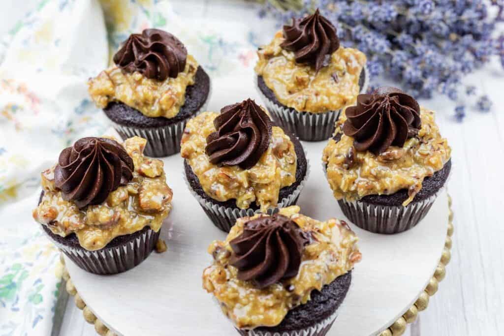 Six chocolate cupcakes topped with a nutty caramel mixture and a swirl of chocolate frosting, arranged on a round tray.