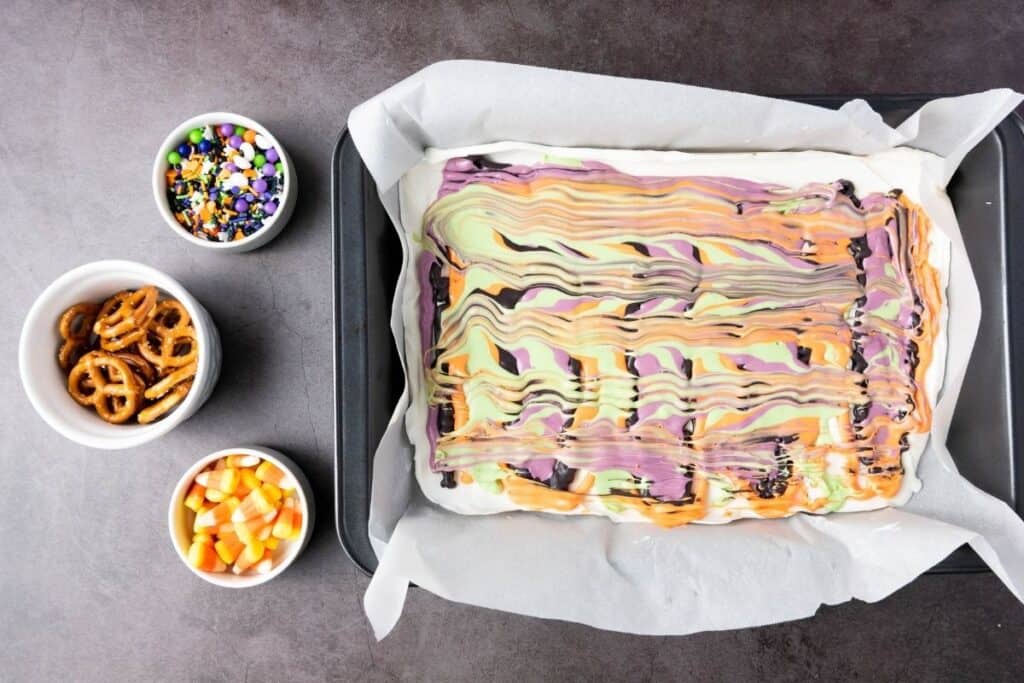 A baking pan with swirled pastel colored bark on parchment paper, next to small bowls of Halloween sprinkles, pretzels, and candy corn on a dark surface.
