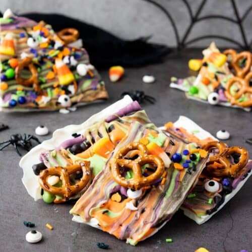Colorful Halloween-themed chocolate bark topped with pretzels, candy eyes, sprinkles, and candy corn, displayed on a gray surface with a spider web decoration in the background.