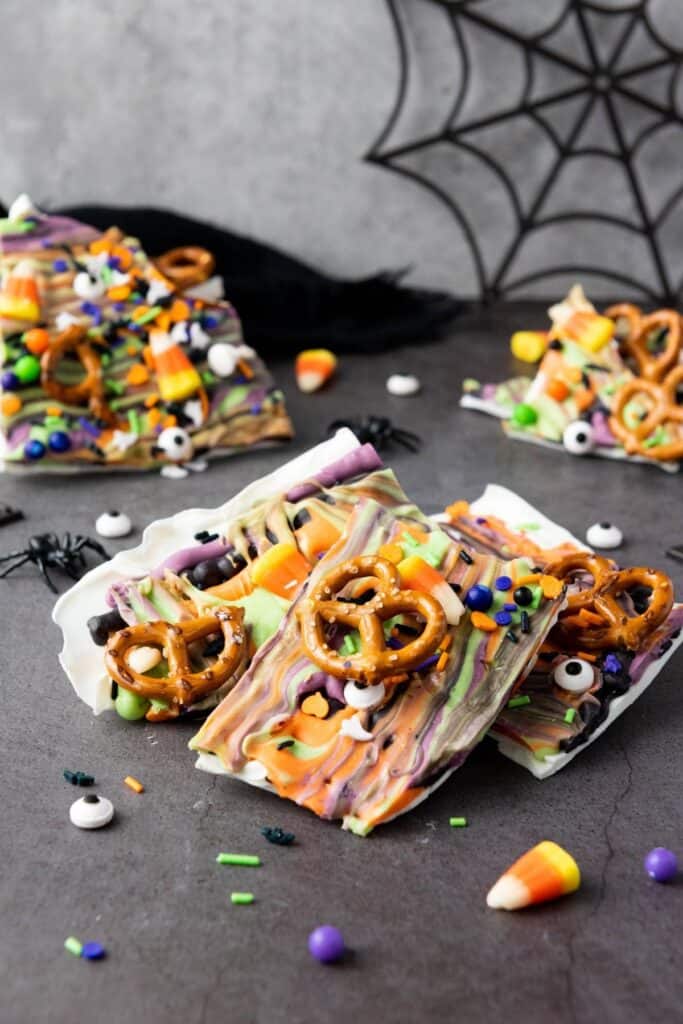 Colorful Halloween-themed chocolate bark topped with pretzels, candy eyes, sprinkles, and candy corn, displayed on a gray surface with a spider web decoration in the background.