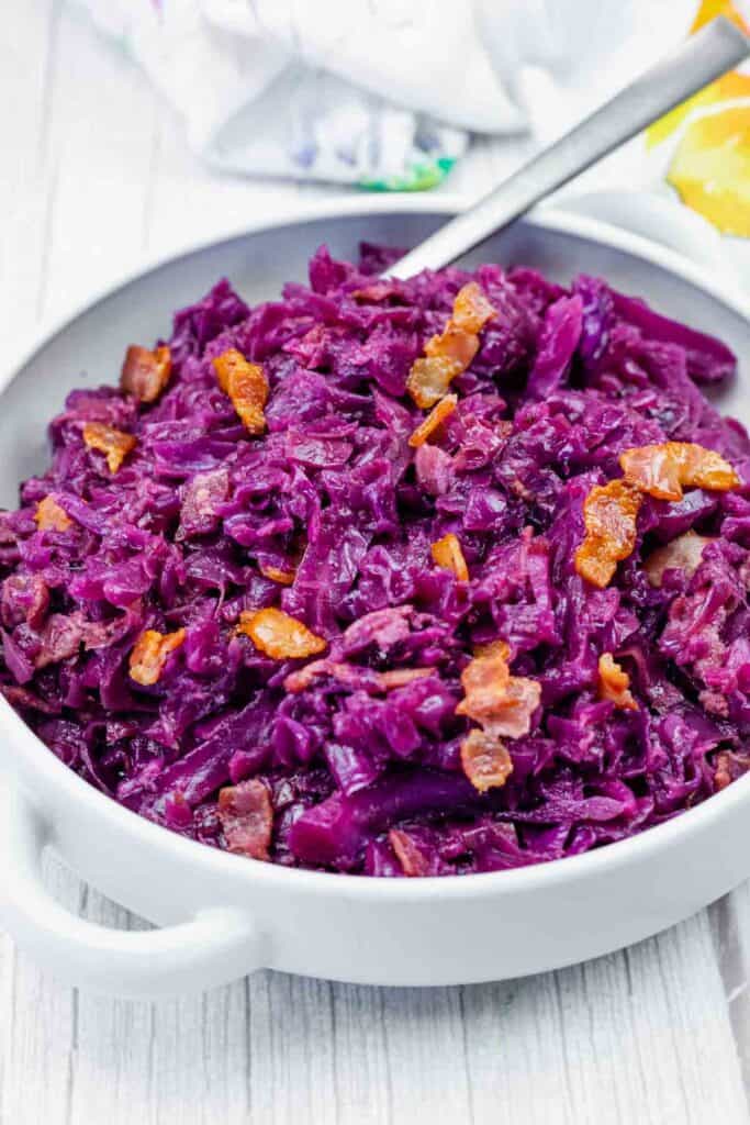 A white bowl filled with cooked red cabbage and bacon pieces, with a serving spoon resting inside.