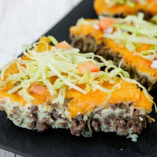 A slice of casserole topped with shredded lettuce, diced tomatoes, ground beef, melted cheese, and a creamy layer, served on a dark plate.