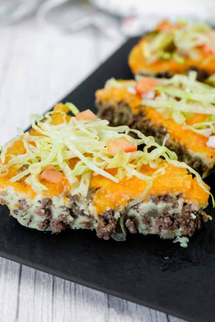 A slice of casserole topped with shredded lettuce, diced tomatoes, ground beef, melted cheese, and a creamy layer, served on a dark plate.