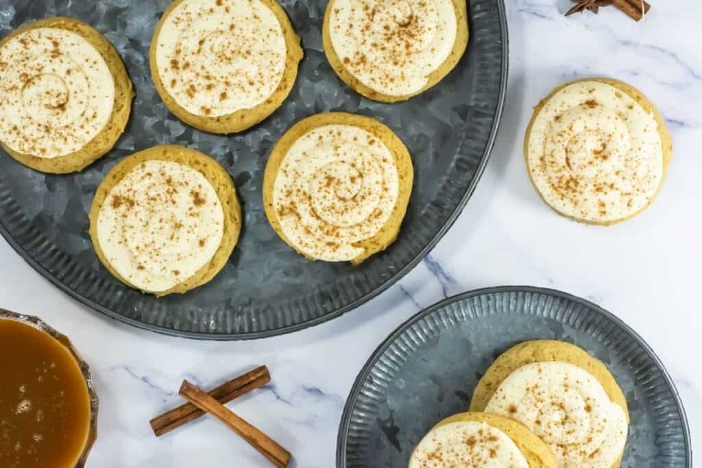 Cookies with white frosting and brown sprinkles are arranged on a metal plate. Decorative autumn items are placed nearby.
