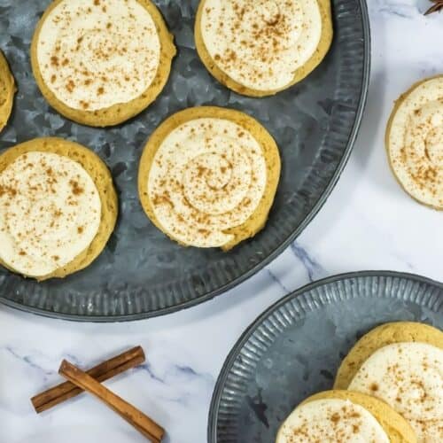 Cookies with white frosting and brown sprinkles are arranged on a metal plate. Decorative autumn items are placed nearby.