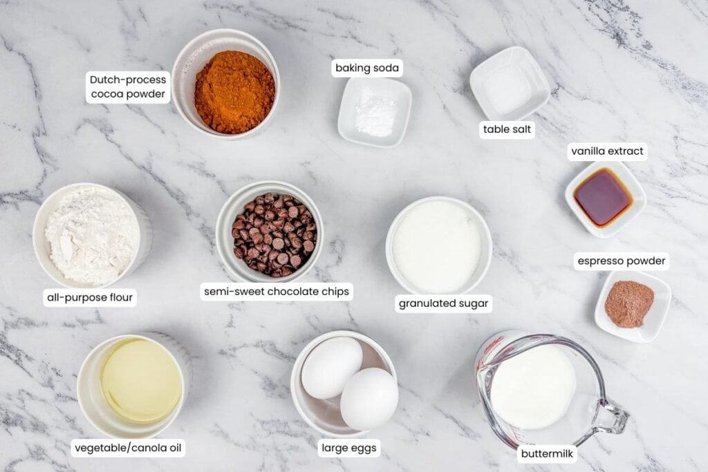 Ingredients for baking are arranged on a marble counter, including cocoa powder, flour, chocolate chips, sugar, eggs, oil, buttermilk, baking soda, salt, vanilla, and espresso powder.