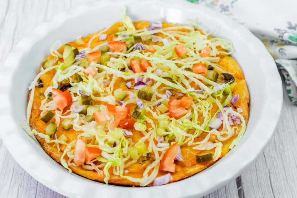 A baked dish topped with shredded lettuce, diced tomatoes, pickles, and red onion sits in a white pie dish on a light wooden surface.