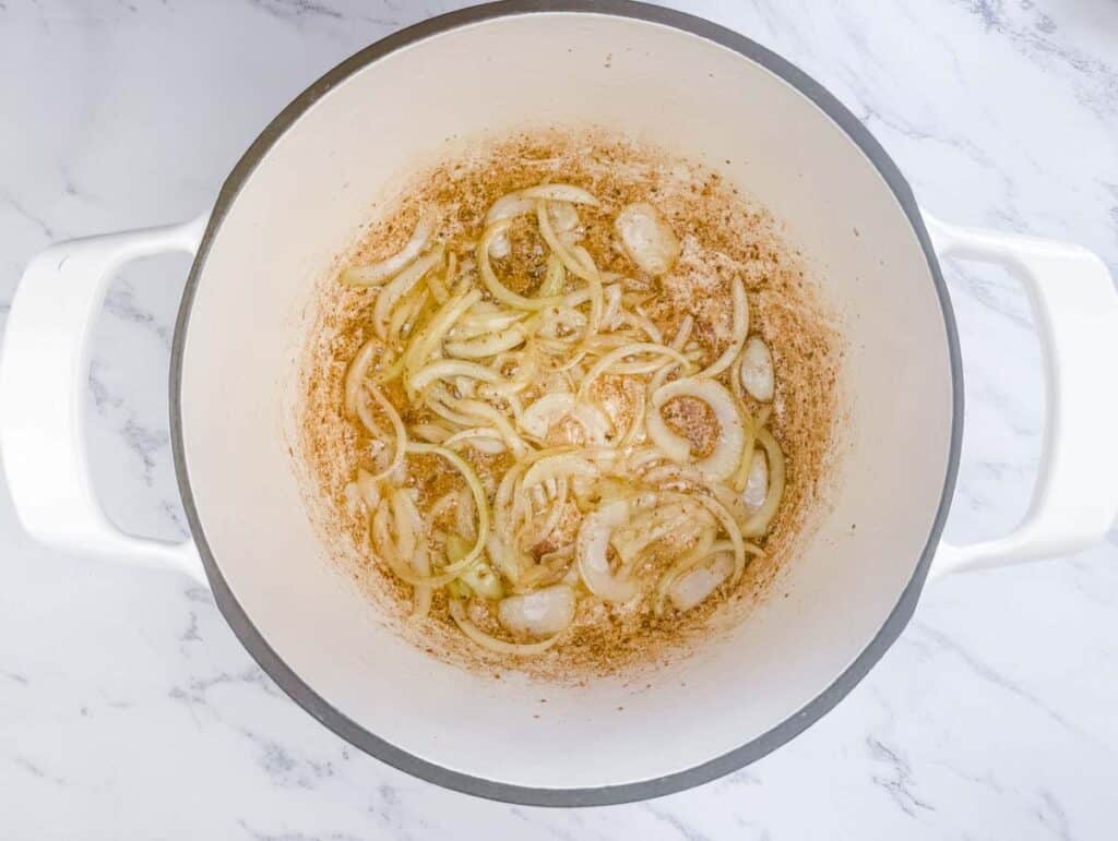 Sliced onions sautéing in a white pot on a marble countertop, with browned bits visible on the bottom of the pot.