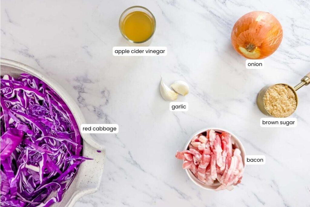 Ingredients on a marble surface: sliced red cabbage, apple cider vinegar, a whole onion, brown sugar, two garlic cloves, and chopped bacon.