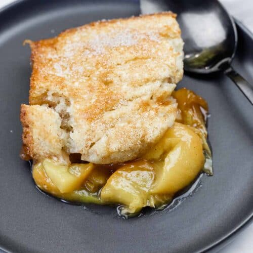 A serving of apple cobbler with a golden-brown crust and syrupy apple filling on a dark plate with a spoon beside it.