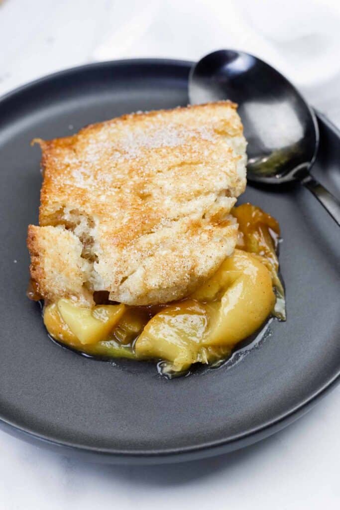A serving of apple cobbler with a golden-brown crust and syrupy apple filling on a dark plate with a spoon beside it.