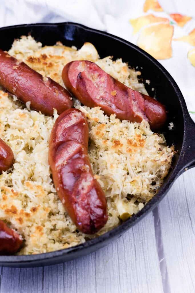 Sausages on a bed of baked sauerkraut in a black cast iron skillet, placed on a light-colored wooden surface.