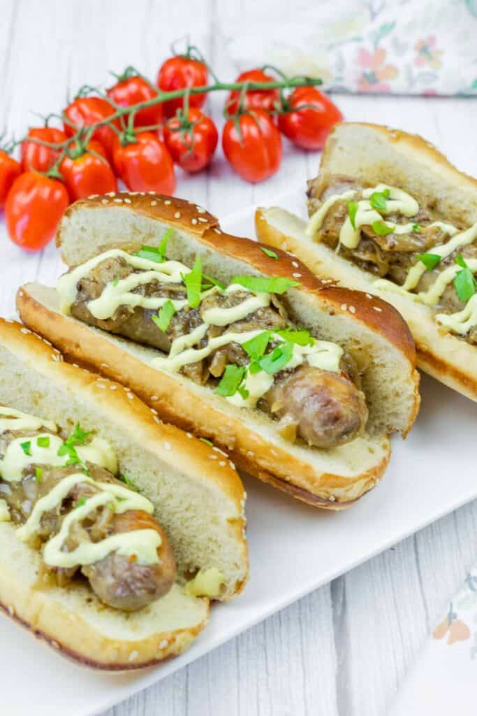 Three sausage sandwiches in buns with mustard, onions, and parsley on a white plate, with a bunch of cherry tomatoes in the background.