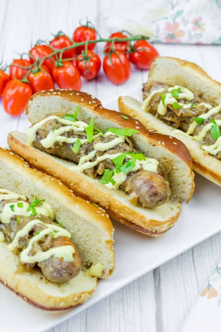 Three sausage sandwiches in buns with mustard, onions, and parsley on a white plate, with a bunch of cherry tomatoes in the background.