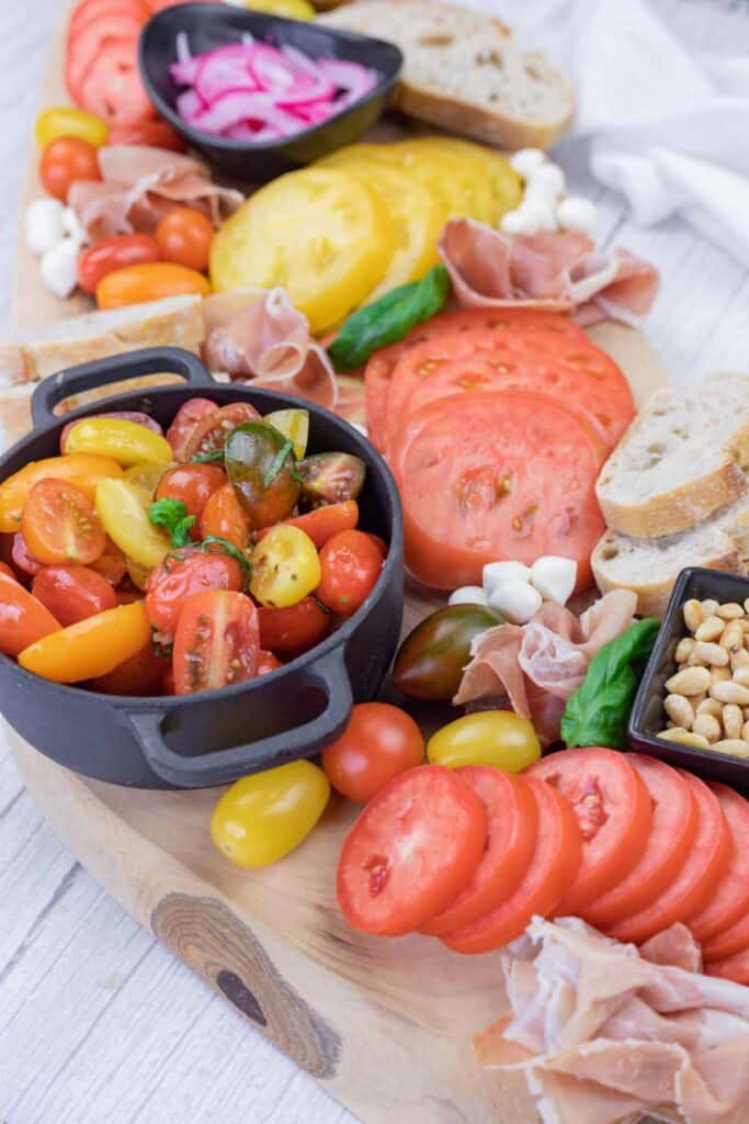A wooden board with sliced tomatoes, cherry tomatoes in a bowl, prosciutto, mozzarella balls, bread, pickled onions, and a small dish of pine nuts.