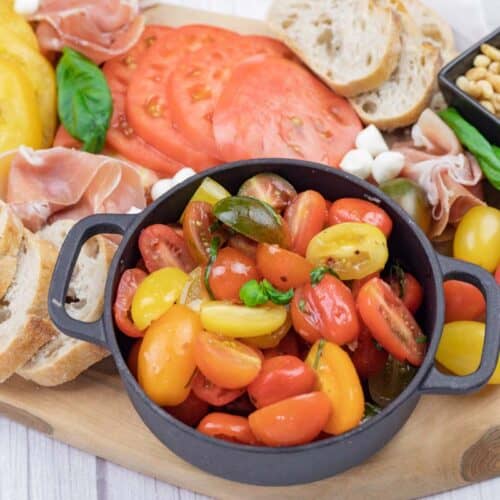 A charcuterie board with sliced tomatoes, bread, cherry tomatoes, prosciutto, mozzarella balls, basil, and a bowl of pine nuts.