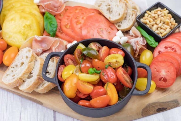 A charcuterie board with sliced tomatoes, bread, cherry tomatoes, prosciutto, mozzarella balls, basil, and a bowl of pine nuts.