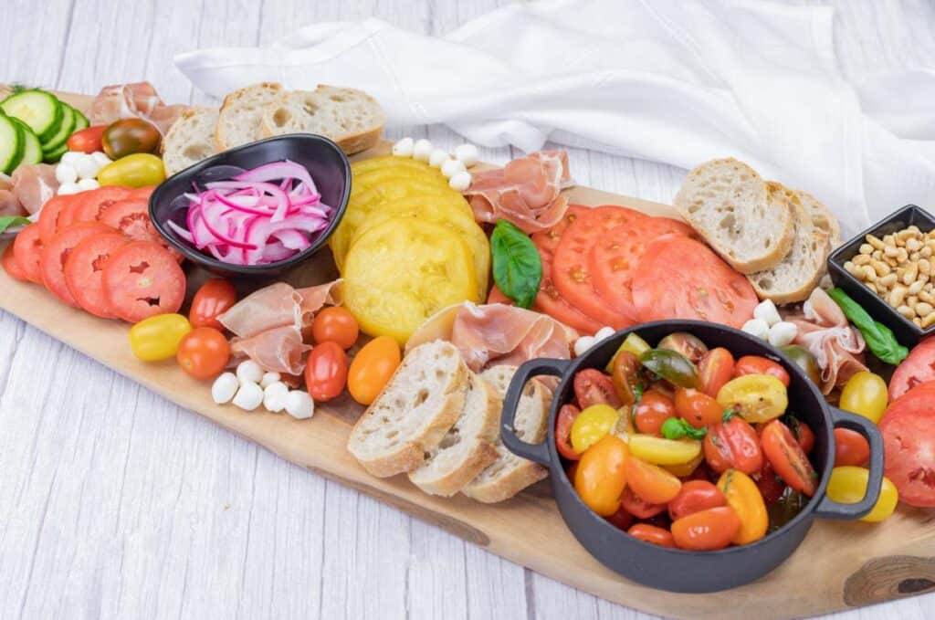 A wooden board with sliced tomatoes, cherry tomatoes, prosciutto, mozzarella balls, pickled onions, bread slices, pine nuts, and a white napkin in the background.