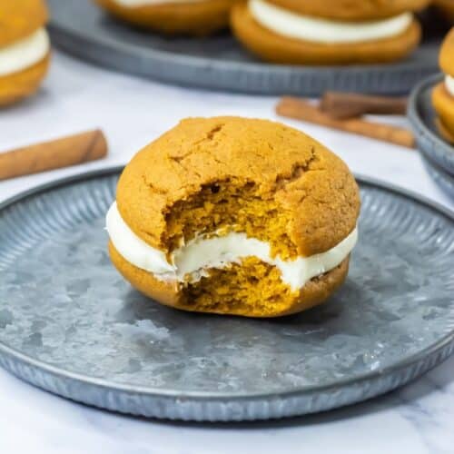 Pumpkin whoopie pie with cream filling on a metal plate, partially eaten to reveal its soft, center. More pies are blurred in the background.