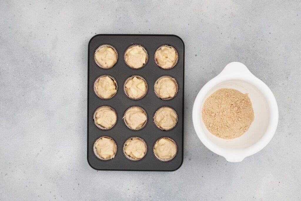 Cream cheese frosting in cupcake liners ready for baking, with a bowl of crushed graham crackers on a gray surface.