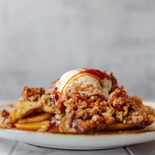 Creamy apple dessert with crumble topping and caramel sauce, topped with vanilla ice cream, served on a white plate.