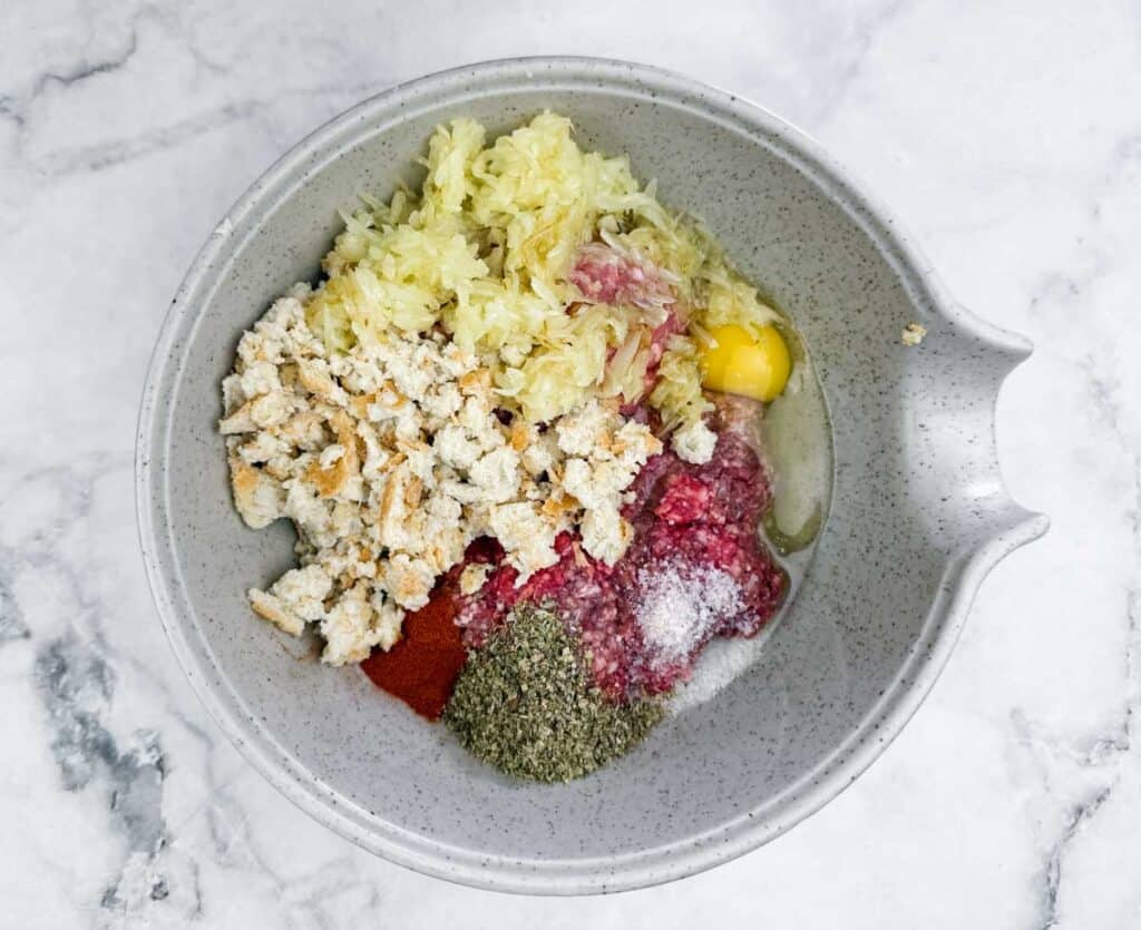 A mixing bowl containing soaked bread, ground beef, ground pork, onion, egg, parsley, mustard, marjoram, salt, pepper, and paprika on a marble surface.