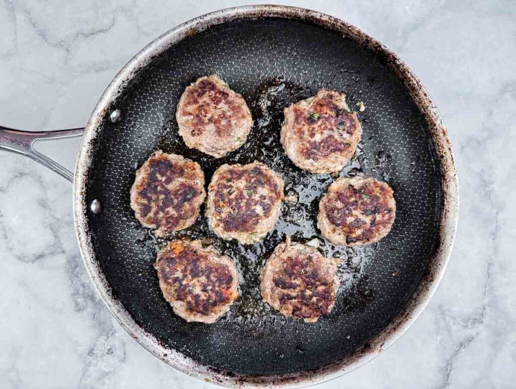 Seven browned patties cooking in a black frying pan on a light gray countertop.