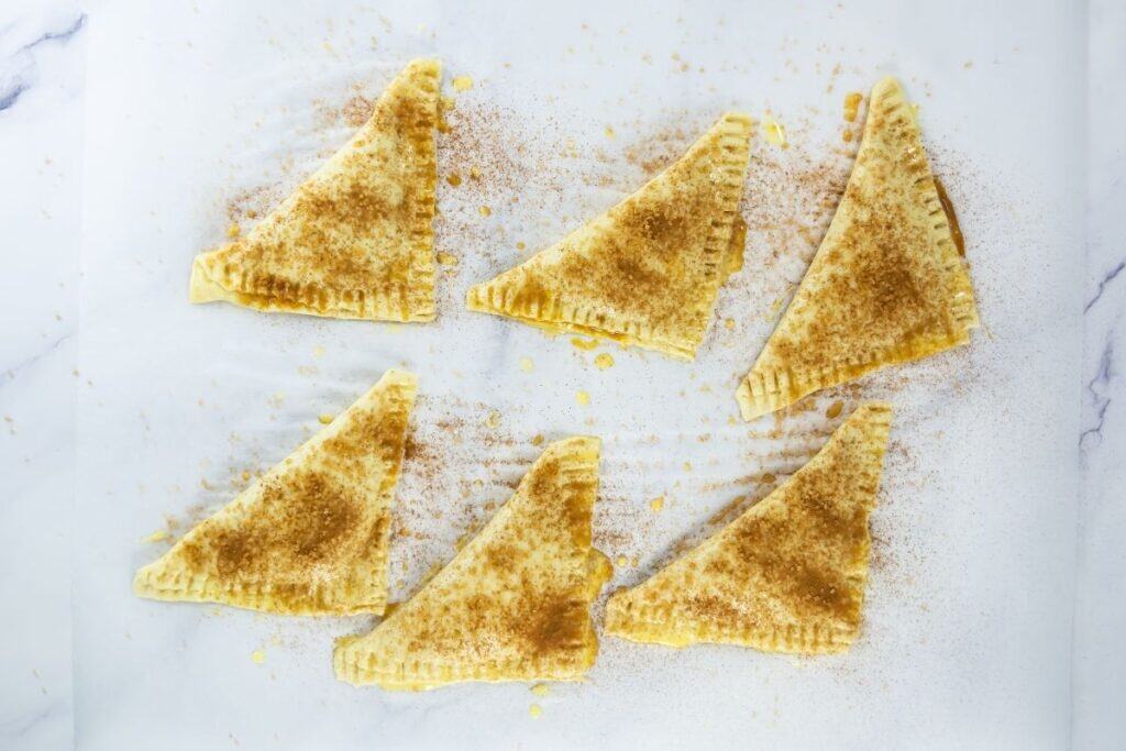 Unbaked pumpkin turnovers on parchment, sprinkled with spices and sealed at the edges.