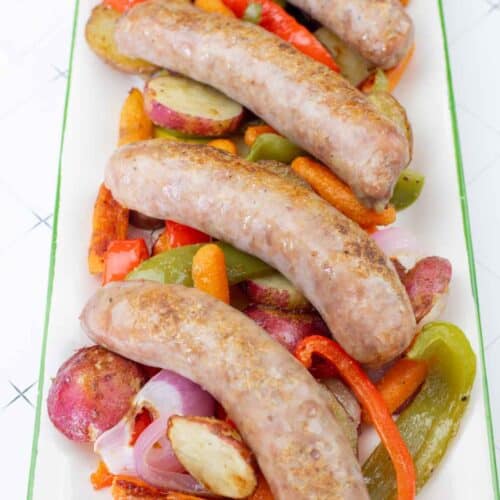 Roasted bratwurst sausages served on a platter with potatoes, carrots, peppers, and onions.