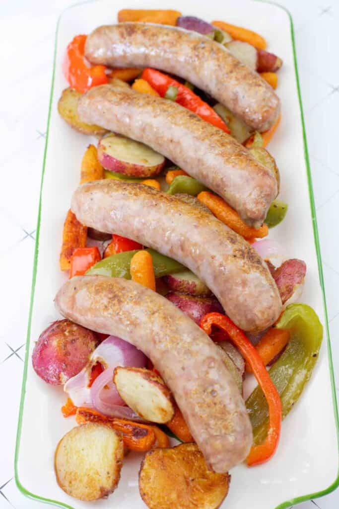 Roasted bratwurst sausages served on a platter with potatoes, carrots, peppers, and onions.