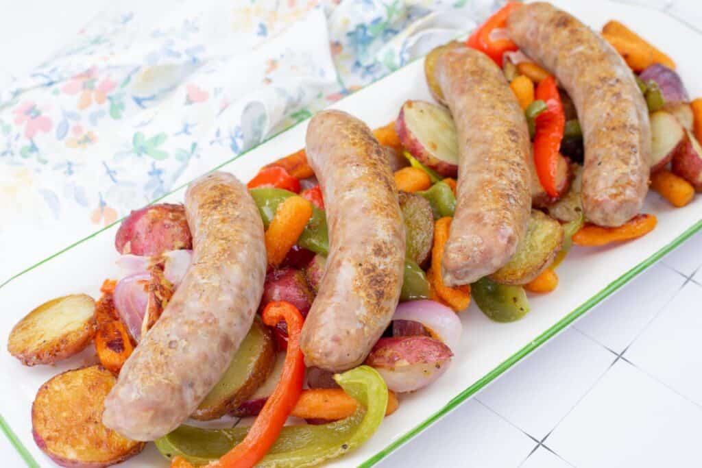 Roasted bratwurst sausages served on a platter with potatoes, carrots, peppers, and onions.