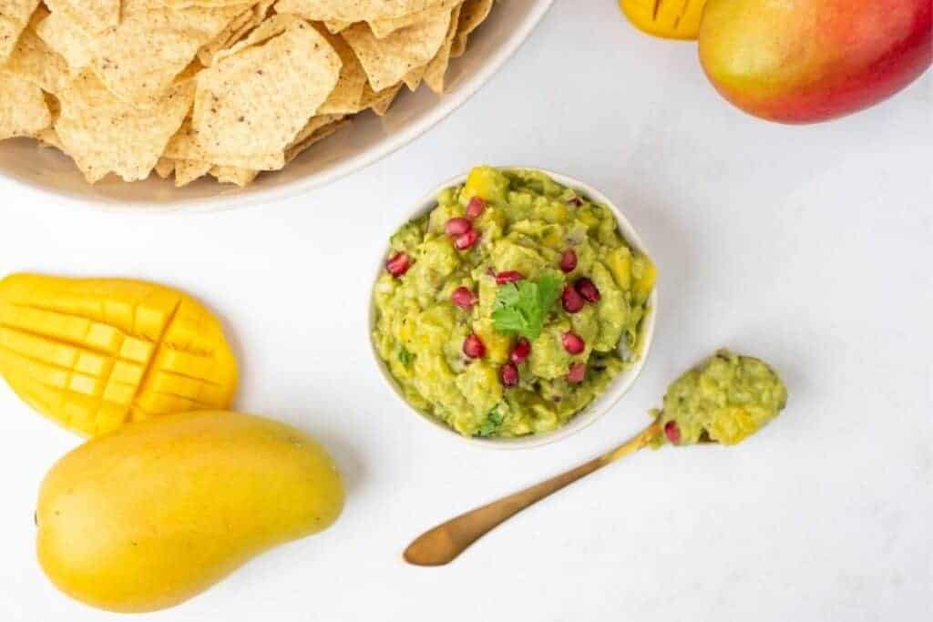 A bowl of tropical mango guacamole surrounded by a bowl of nachos and mango.