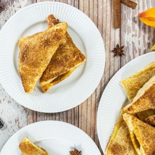 Three white plates with pumpkin turnovers topped with cinnamon sugar, on a rustic wooden table with utensils and autumn-themed decorations.