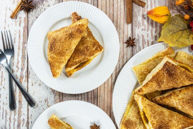 Three white plates with pumpkin turnovers topped with cinnamon sugar, on a rustic wooden table with utensils and autumn-themed decorations.