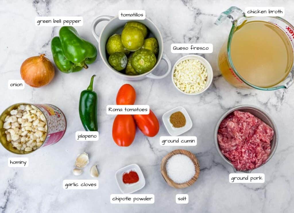 Ingredients for a recipe are arranged on a marble surface, including ground pork, hominy, green bell pepper, tomatillos, Roma tomatoes, onion, jalape&ntilde;o, garlic, cheese, spices, and chicken broth.