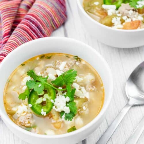 Bowls of Green Pork Posole garnished with cilantro, jalape&ntilde;o slices, and crumbled cheese sit on a light wooden surface.