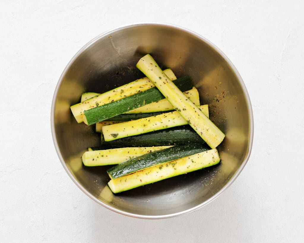 Fresh zucchini sticks seasoned with herbs in a stainless steel bowl for healthy snacks.