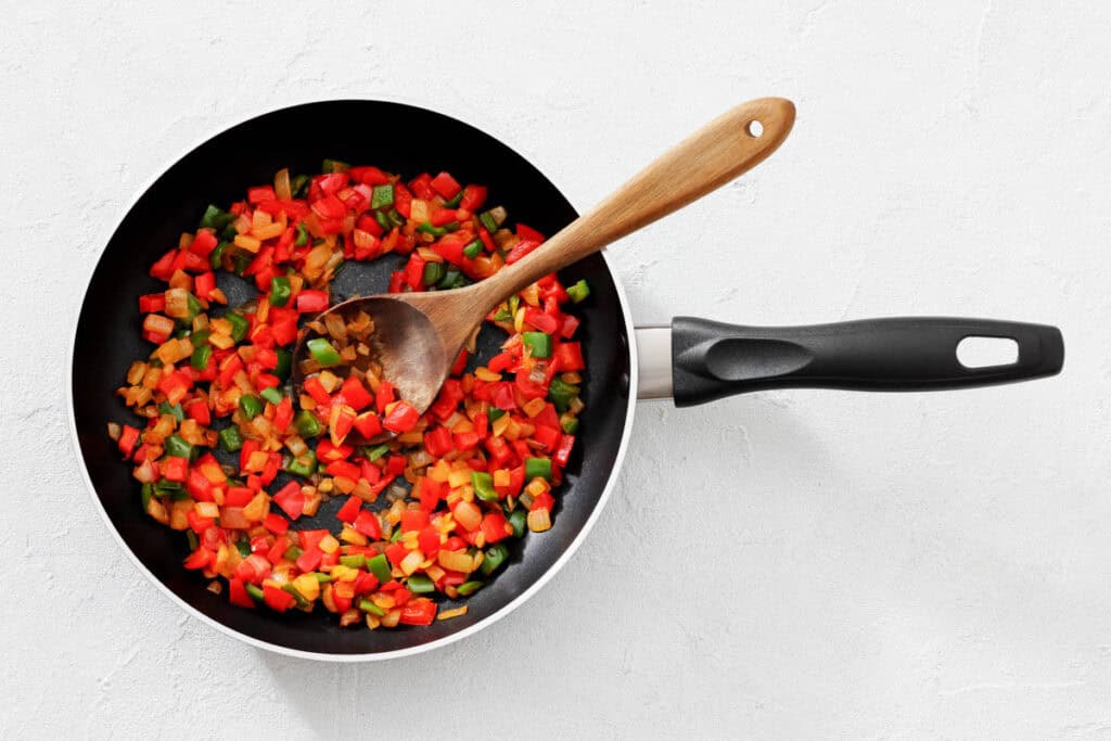 Diced colorful vegetables cooking in a black frying pan with a wooden spoon on a white surface.
