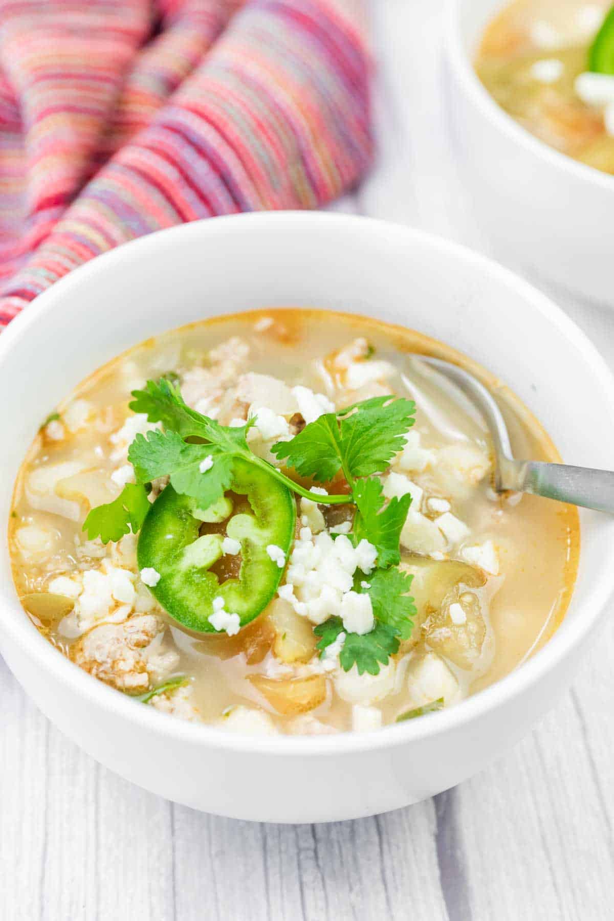 A bowl of Green Pork Posole topped with sliced jalape&ntilde;o, cilantro, and crumbled cheese, with a spoon in the bowl and a striped cloth in the background.