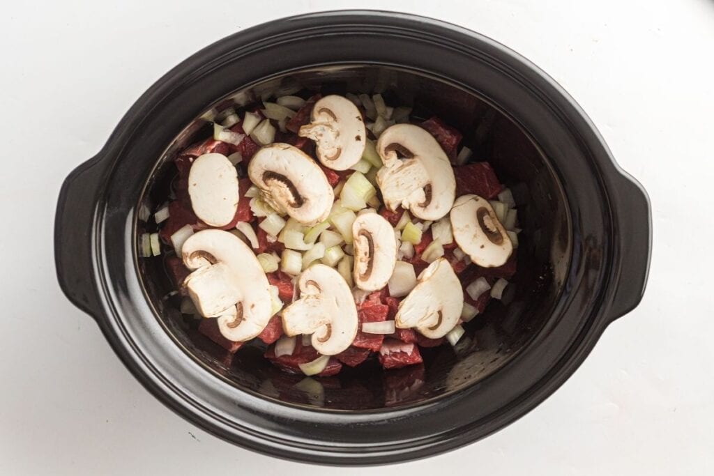 Image of beef stew ingredients in a slow cooker, featuring chunks of beef, sliced mushrooms, and chopped onions.