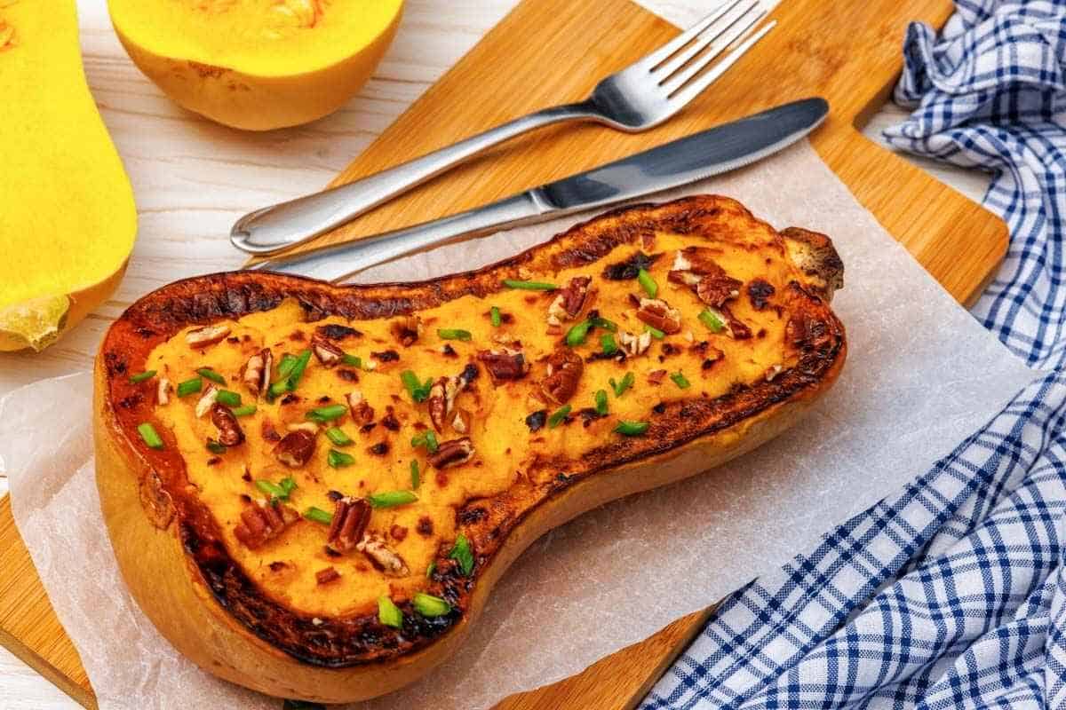 A baked Butternut Squash Mash with Parmesan, garnished with chopped nuts and herbs, served on parchment paper, accompanied by a fork, knife, and a blue checkered cloth.