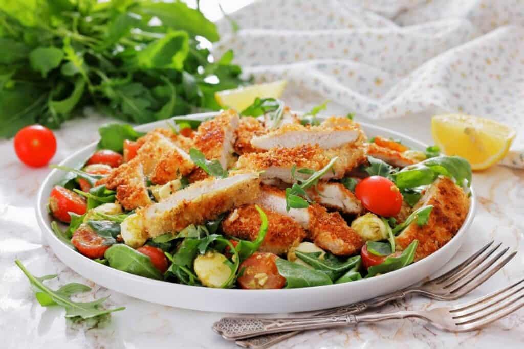 Chicken Parmesan Salad is arranged on a clean white plate, making all the colors stand out. Crispy, golden chicken cutlets are sliced and layered over fresh arugula, with cherry tomatoes and creamy bocconcini scattered around.
