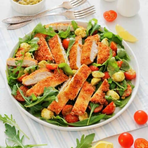 A white plate is filled with a vibrant Chicken Parmesan Salad. Crispy, golden chicken cutlets rest on top of fresh arugula, with cherry tomatoes and creamy bocconcini scattered around.